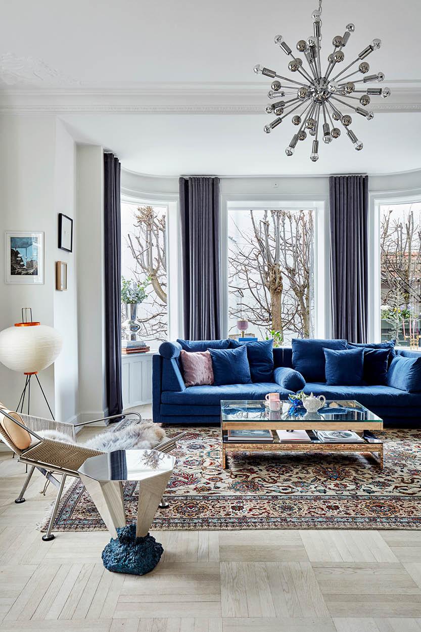 In the living room, curtains from &Drape frame the curved windows. A majestic blue velvet Eilersen sofa is flanked by a Flagline chair by Hans Wegner, Diamond table by FOS, and a rice paper lamp by Isamu Noguchi for Vitra