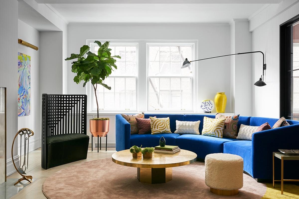 A circular sofa custom-designed by MKCA sites in the living room, upholstered in a bright blue synthetic textile from Naharam. Poufs upholstered in mohair surround a vintage brass table. Far left, a sculptural chair by Charles Rennie Mackintosh. The rug is custom-made by MKCA featuring two forms of broadloom synthetic silk, joined together. (Photo: Brooke Holm)